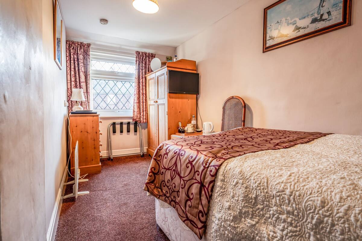 A cozy bedroom with a bed, a wooden wardrobe, a radiator, a window with curtains, and a painting on the wall.