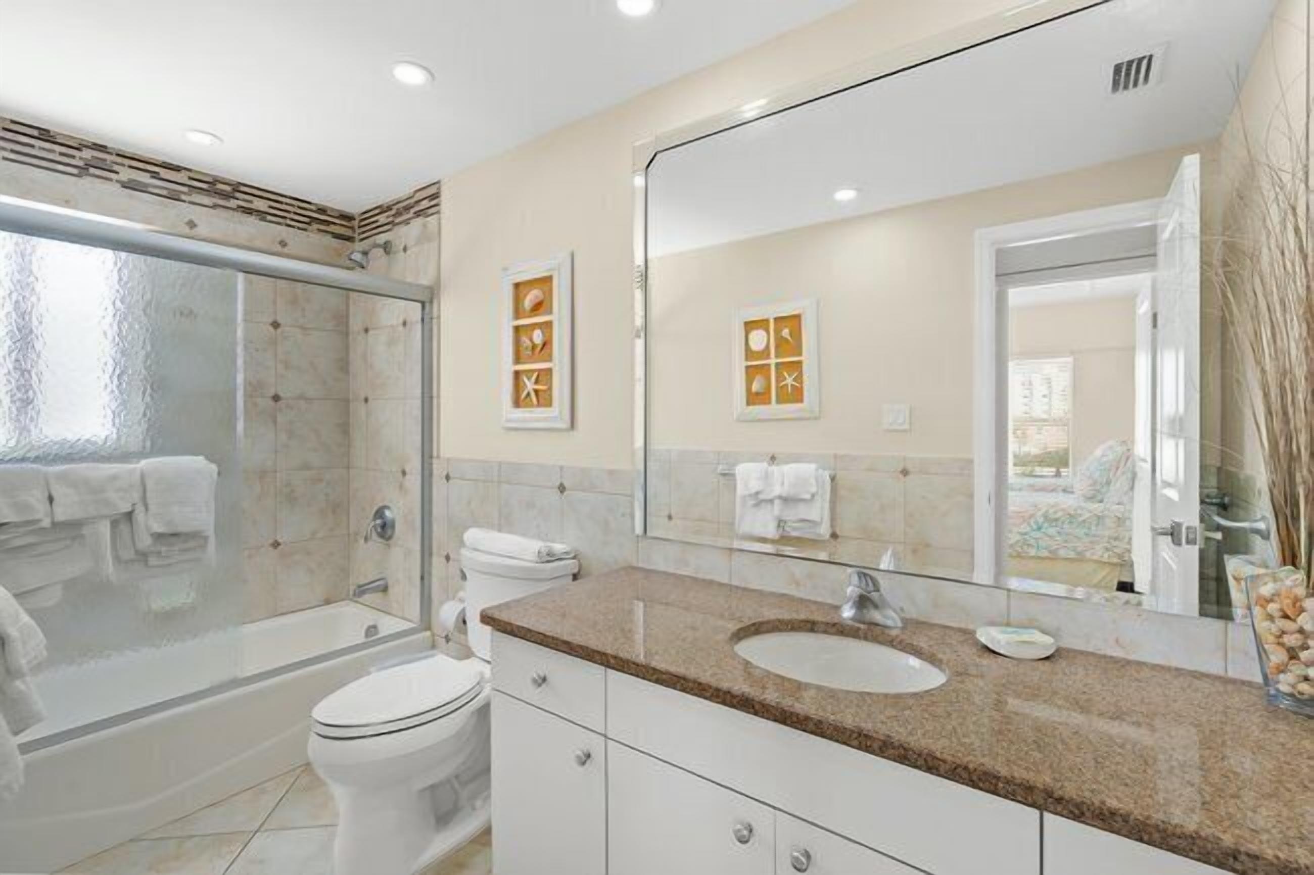 A modern bathroom with a glass shower enclosure, a white toilet, a granite countertop sink, and a large mirror.