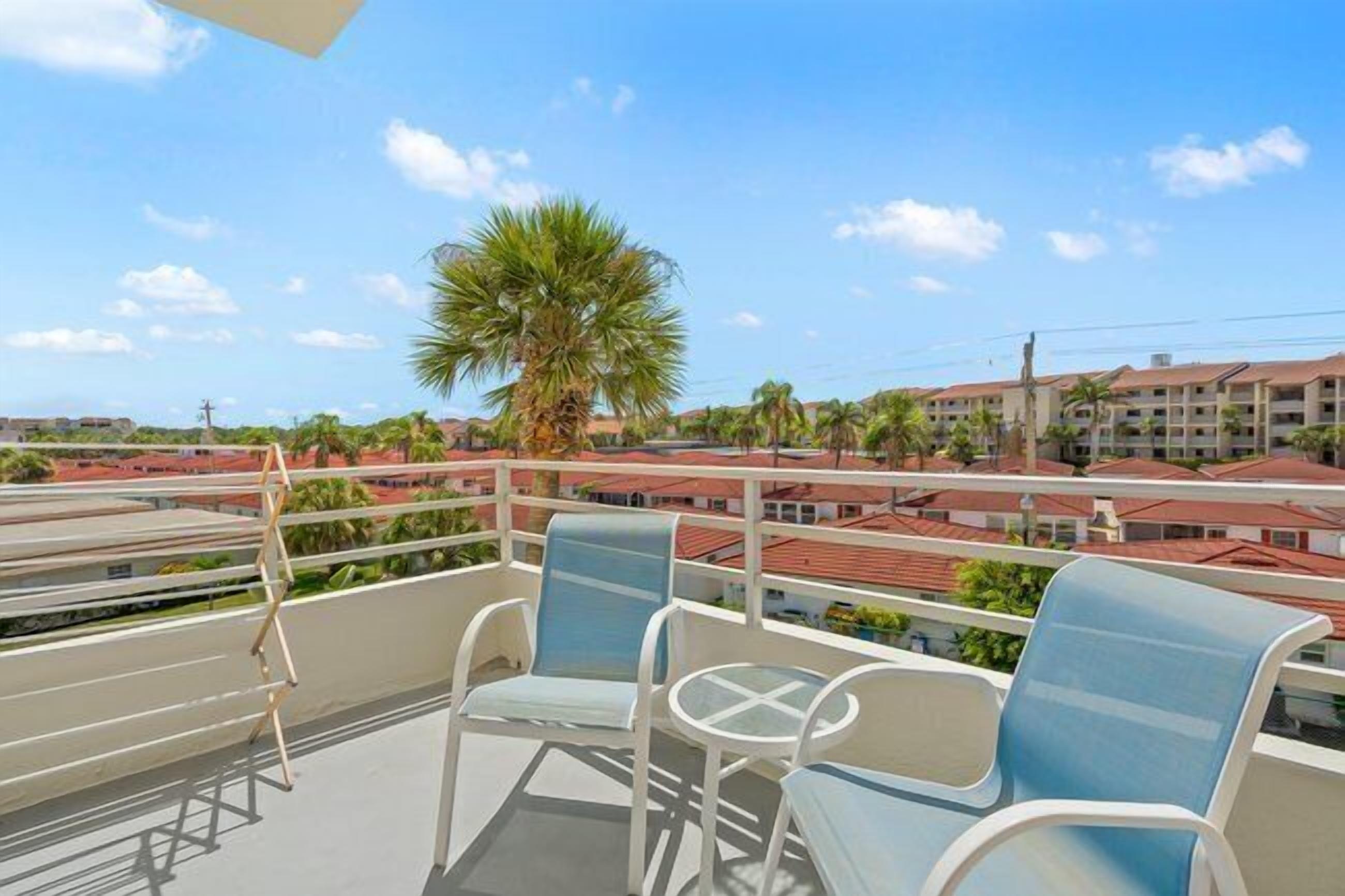 A balcony with two chairs and a small table, overlooking a residential area with red-roofed buildings and palm trees.