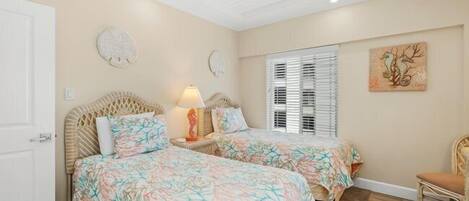 Un dormitorio con dos camas, una mesita de noche con lámpara, una silla y un ventanal con persianas.
