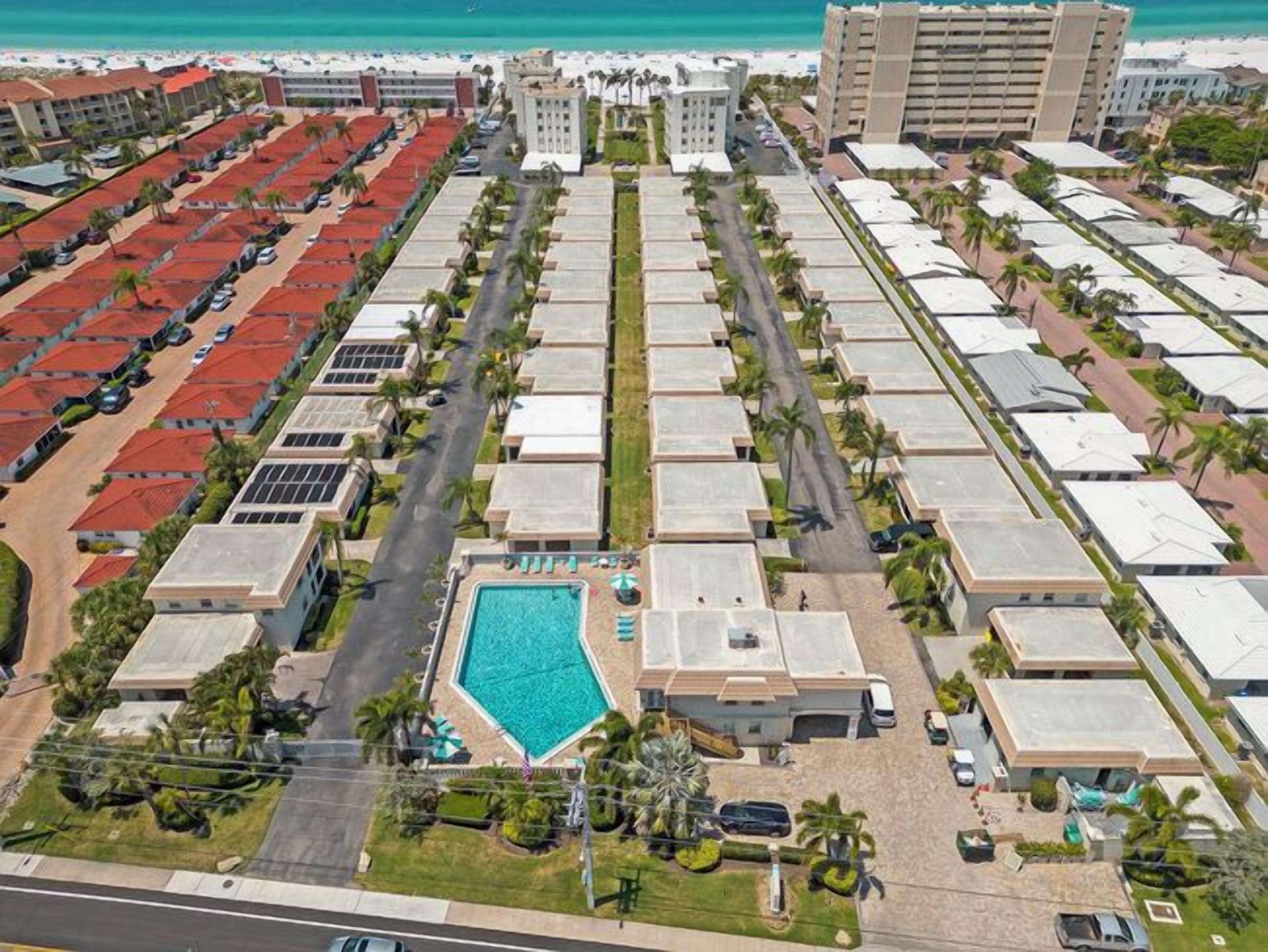 An aerial view of a hotel with multiple buildings, a central swimming pool, and a beach in the background.