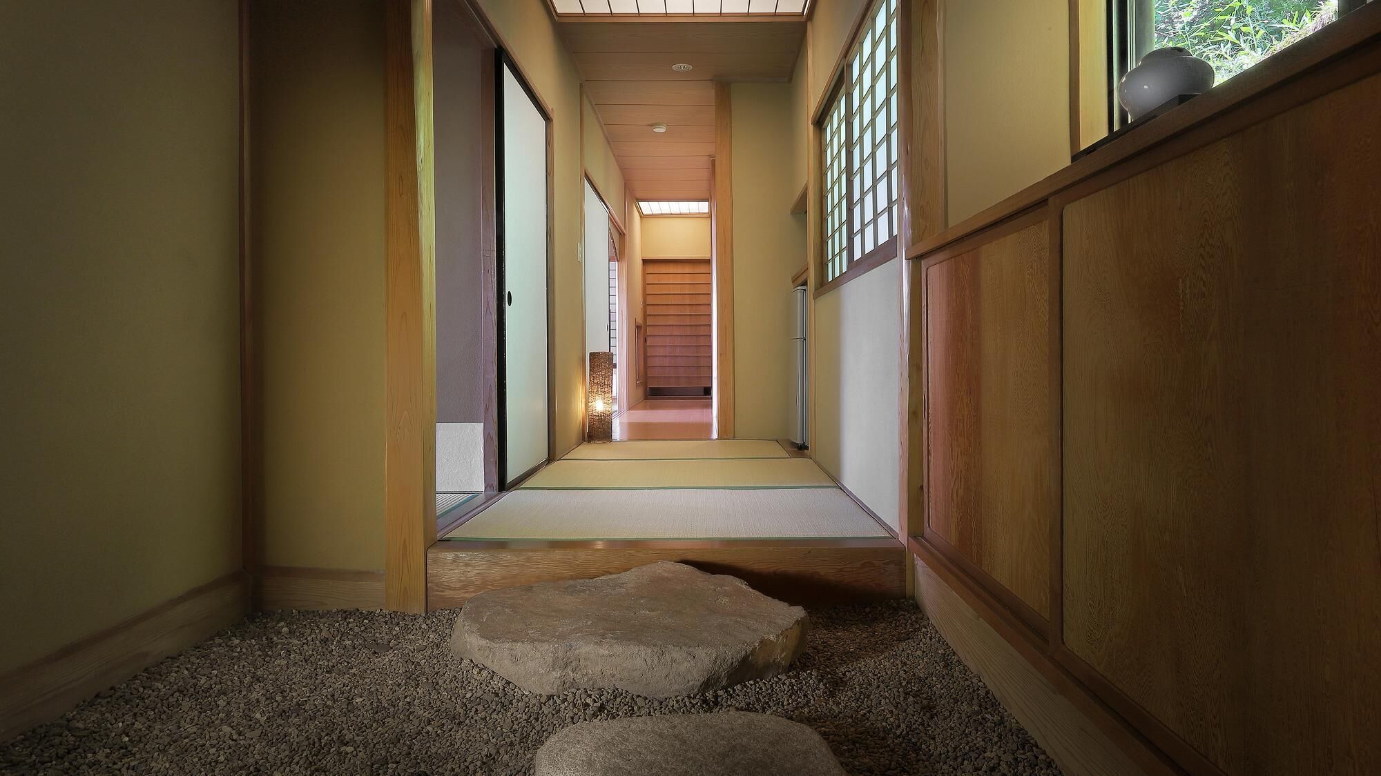 Un pasillo estrecho con piso de madera, un suelo de tatami y un sendero de piedra que conduce a una habitación con puerta corredera.