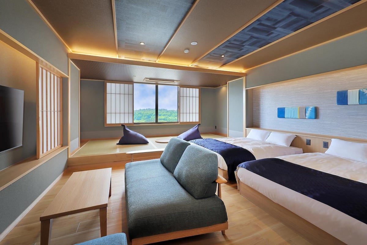 A hotel room with a sofa, two beds, a TV, tatami mats, and a view of greenery.