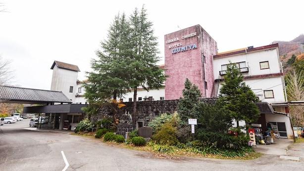 A hotel with a prominent pink tower, a parking area, and a stone wall with greenery. - Ryokan Obara Onsen Hotel Izumiya (Shiroishi)