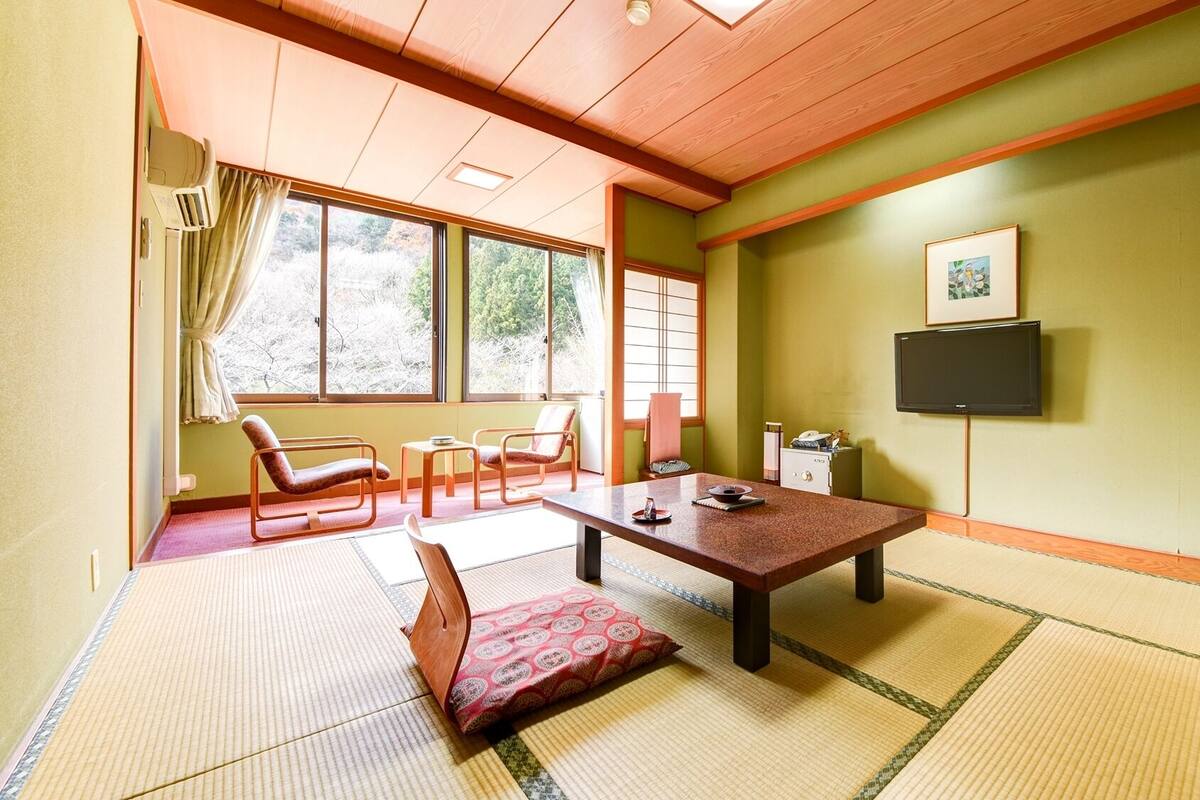 Ein Zimmer mit Tatami-Matten, einem niedrigen Tisch, Stühlen, einem Fernseher und einem Vorhangfenster.