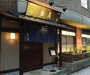A restaurant with a traditional Asian architectural style, featuring a dark wooden entrance, a signboard with Chinese characters, and a small outdoor seating area with plants.