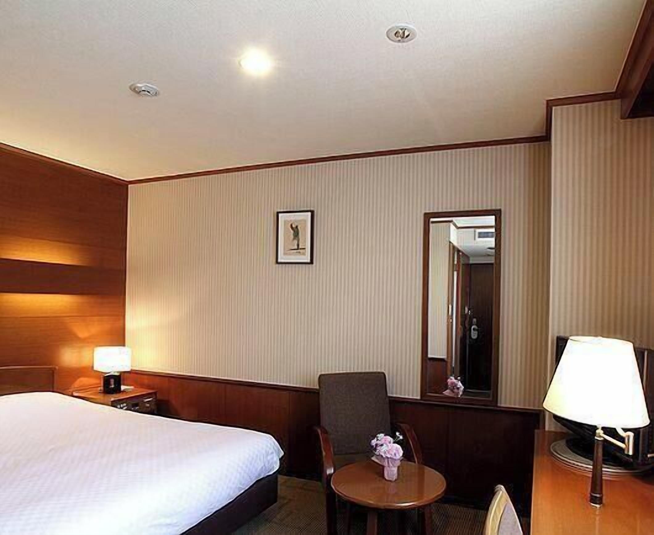 A hotel room with a bed, a chair, a small table, a lamp, a picture on the wall, and a mirror.