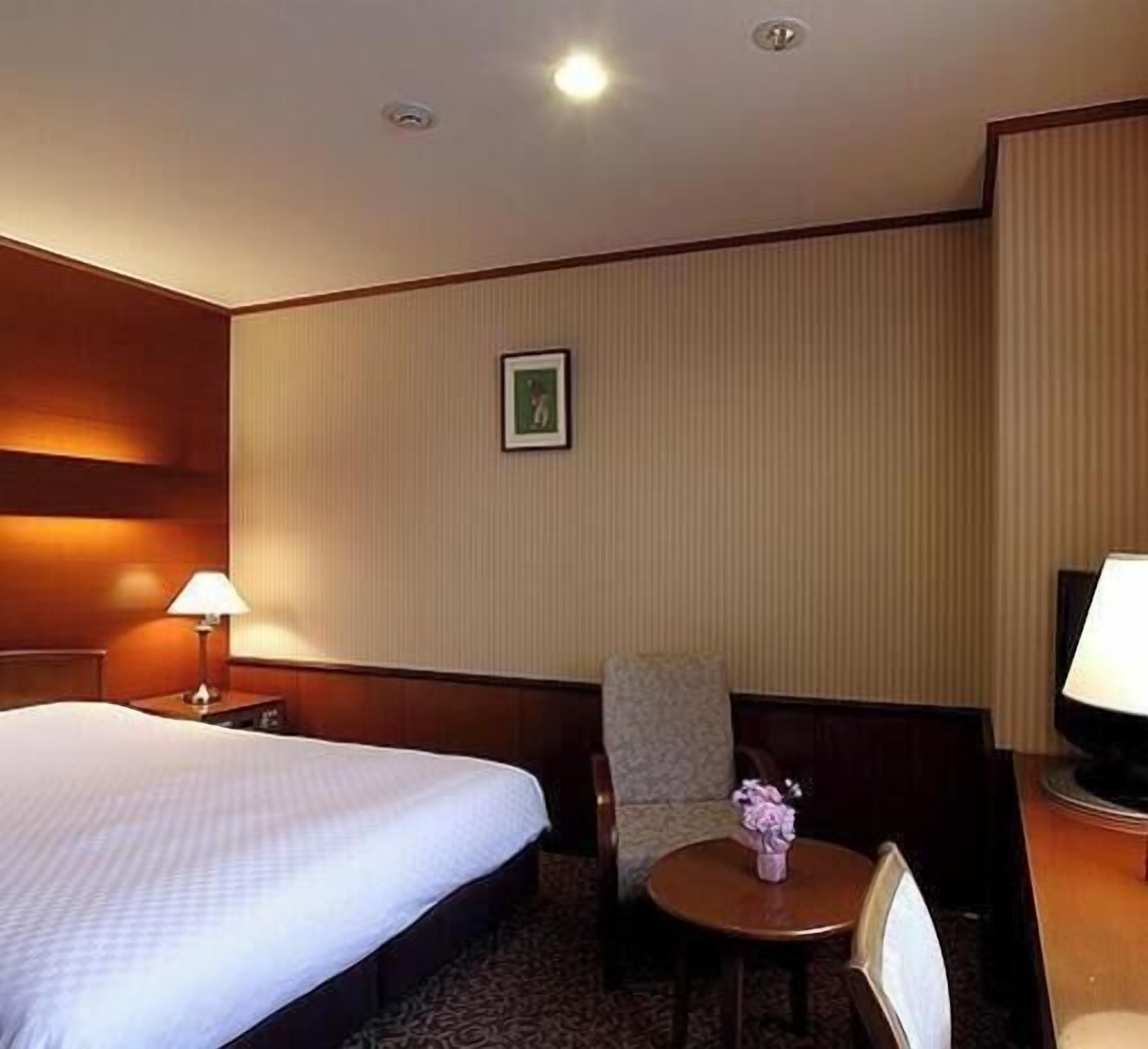 A hotel room with a bed, a chair, a small table with a vase, and a picture on the wall.