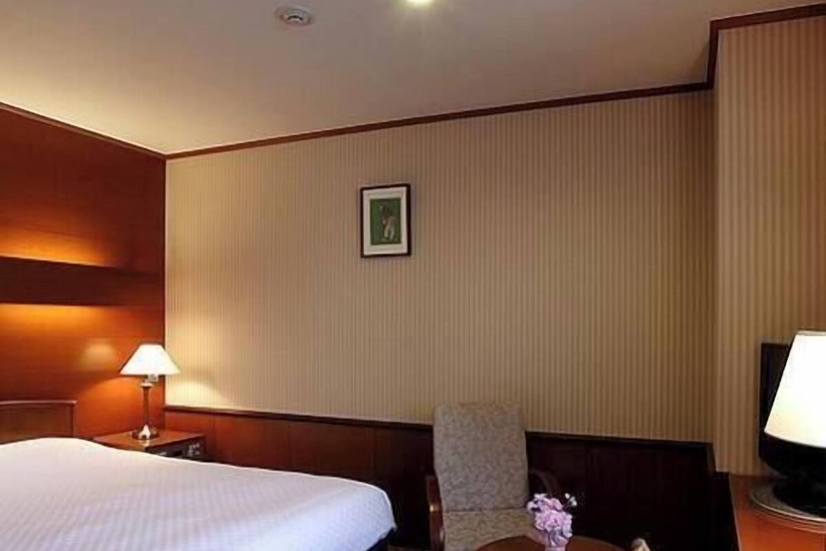 A hotel room with a bed, a chair, a small table with a vase, and a picture on the wall.