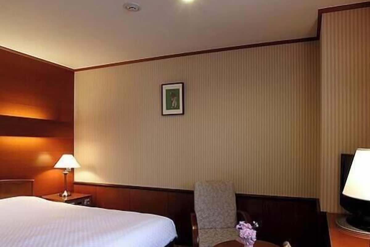 A hotel room with a bed, a chair, a small table with a vase, and a picture on the wall.