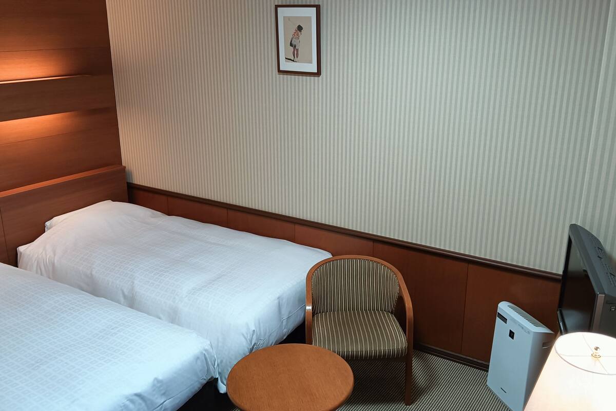 A hotel room with two beds, a chair, a desk, a lamp, and a television.
