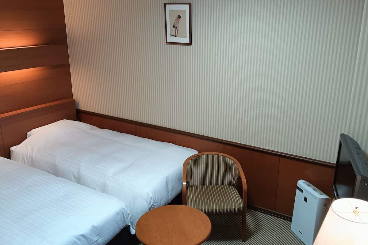 A hotel room with two beds, a chair, a desk, a lamp, and a television.