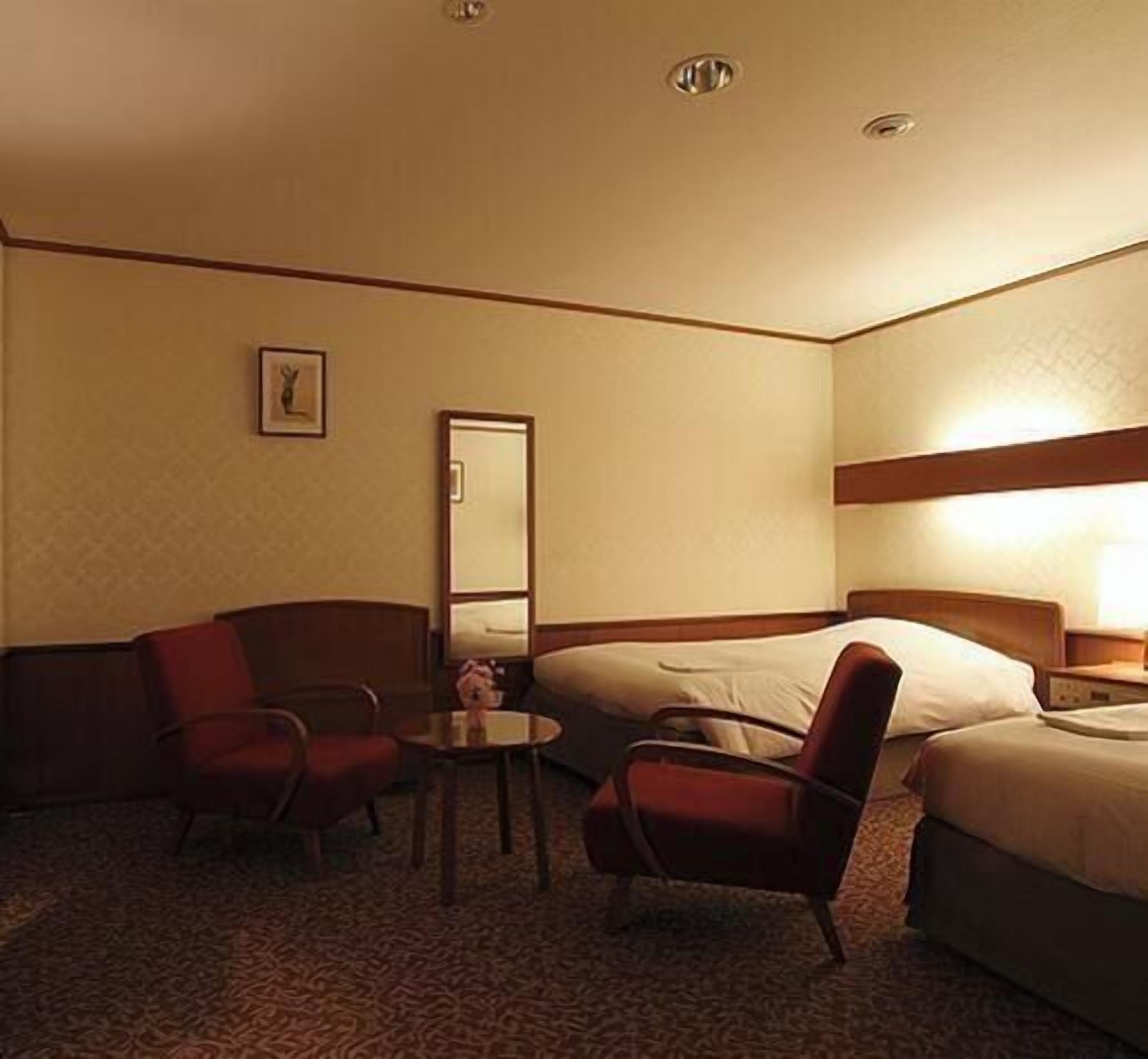A hotel room with two beds, a small table, two armchairs, a mirror, and a picture on the wall.
