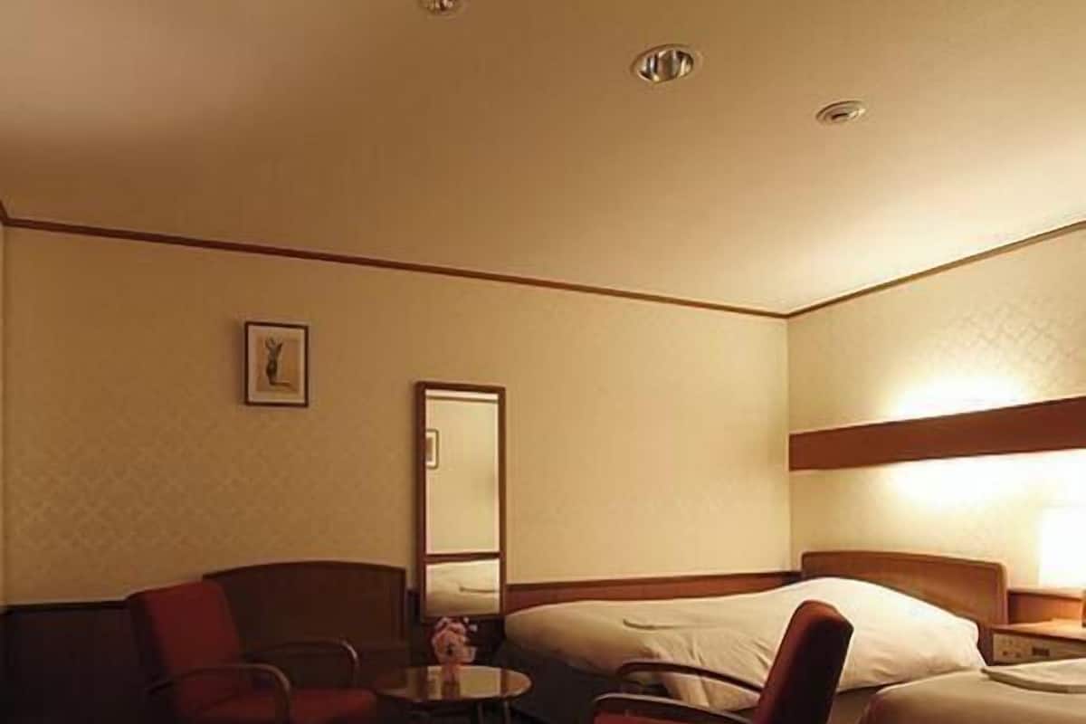 A hotel room with two beds, a small table, two armchairs, a mirror, and a picture on the wall.