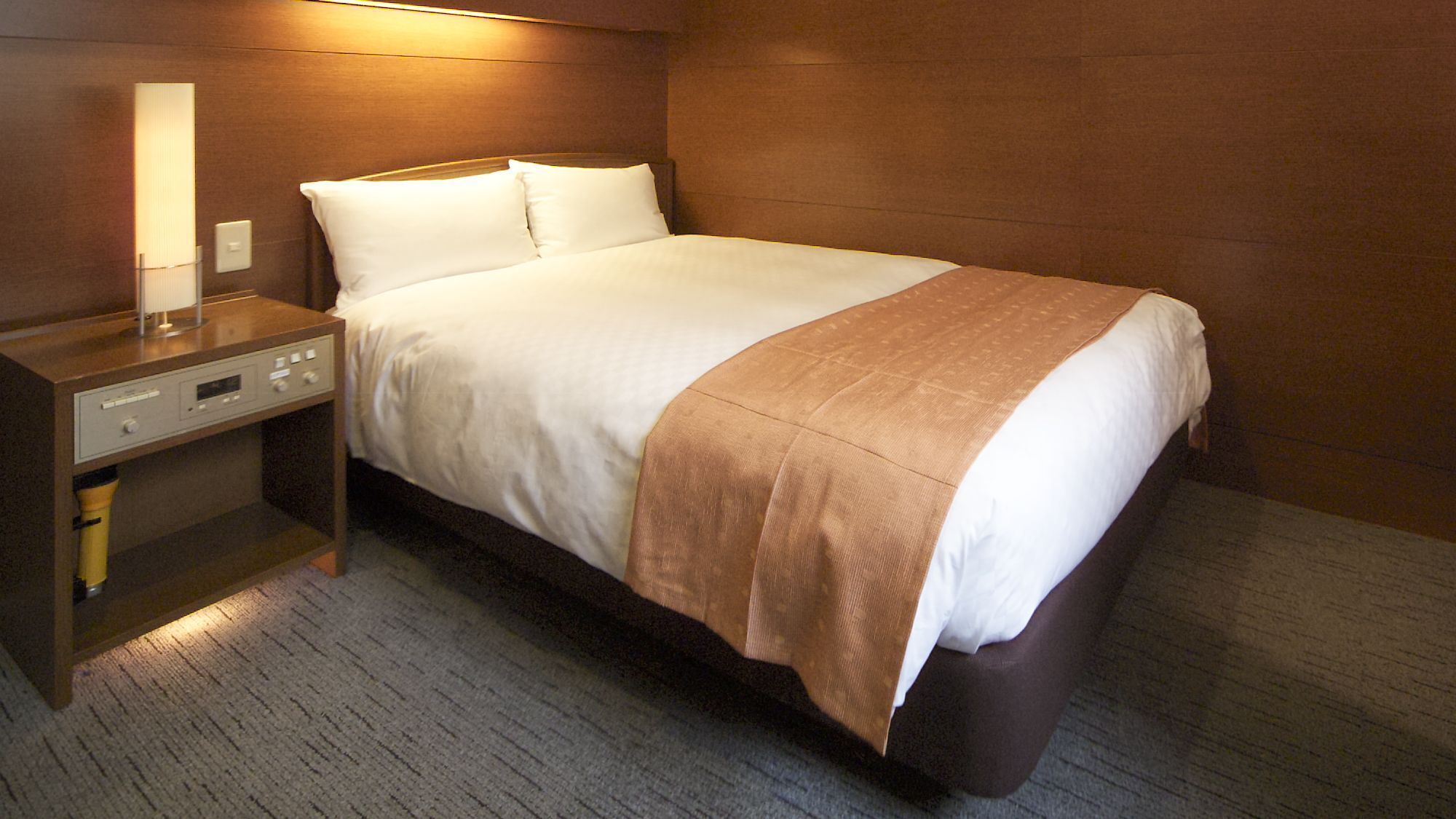 A hotel room with a bed, a nightstand, a lamp, and a control panel.