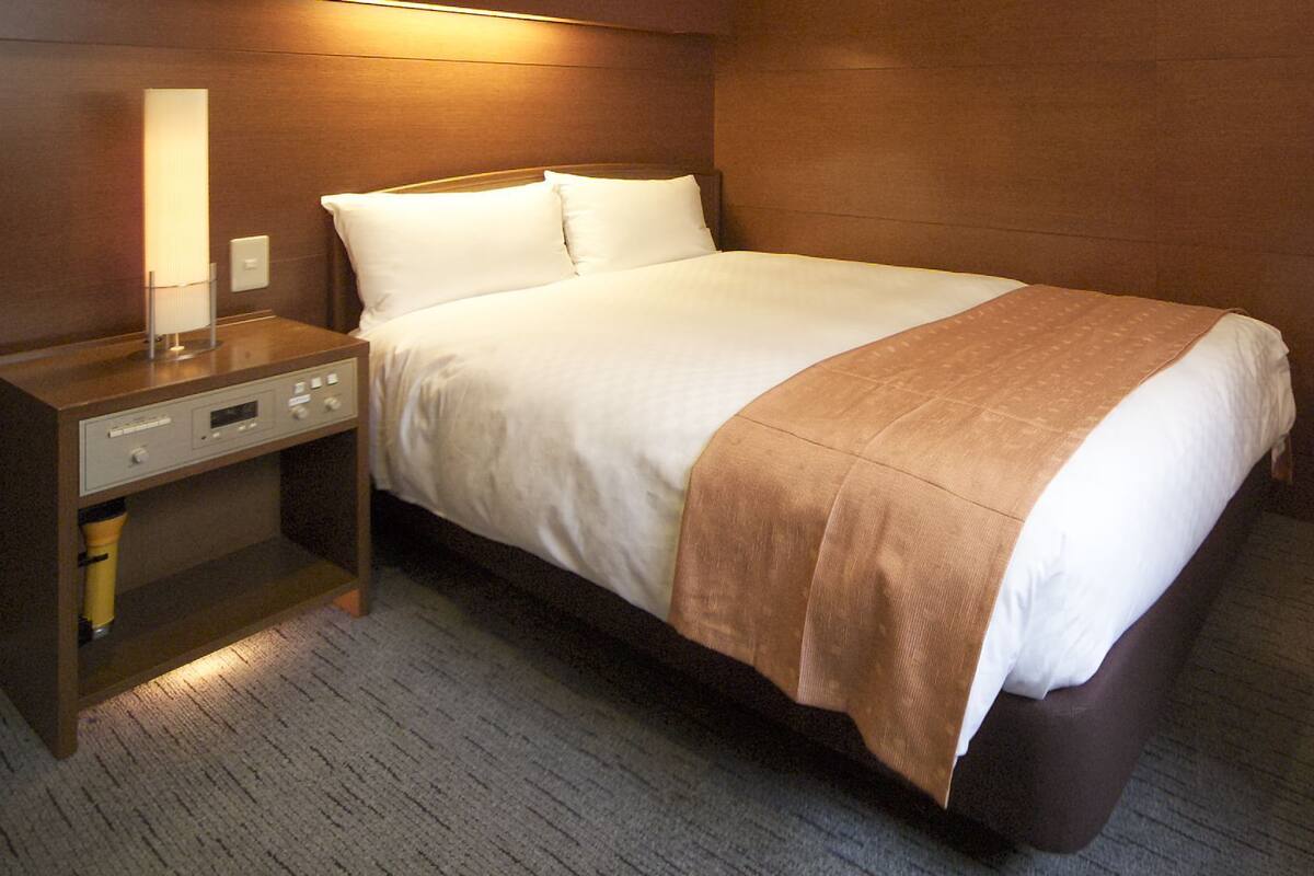 A hotel room with a bed, a nightstand, a lamp, and a control panel.