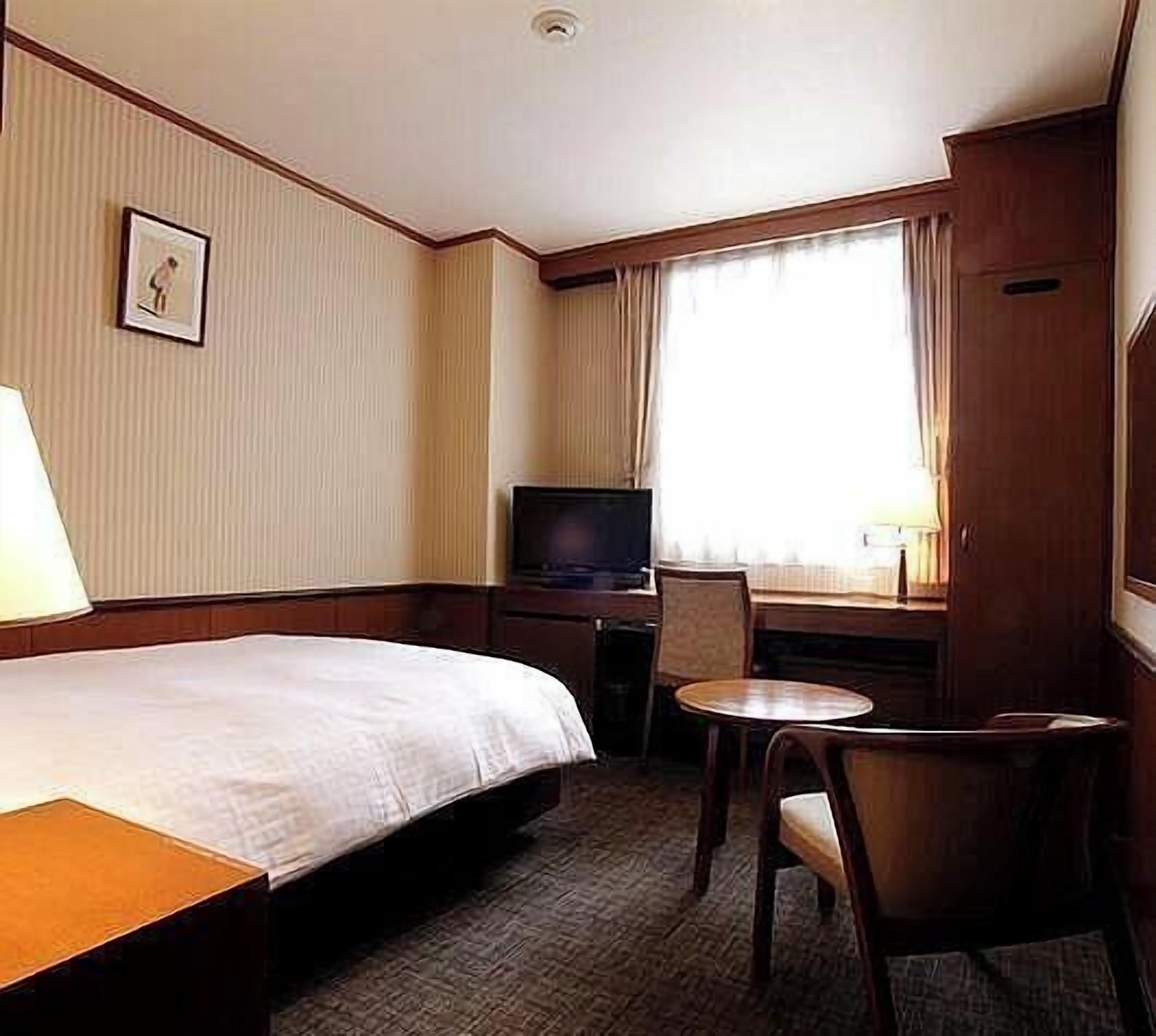 A hotel room with a bed, a desk with a television, a chair, and a small table.