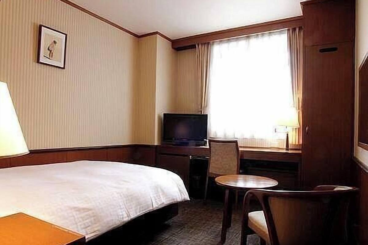 A hotel room with a bed, a desk with a television, a chair, and a small table.