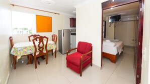 A room with a dining table, chairs, a red armchair, a refrigerator, a bed, and a TV. - 1 Bedroom Apartment en Corales del Sur a 15 Minutos del Aeropuerto (Los Corales Del Sur)