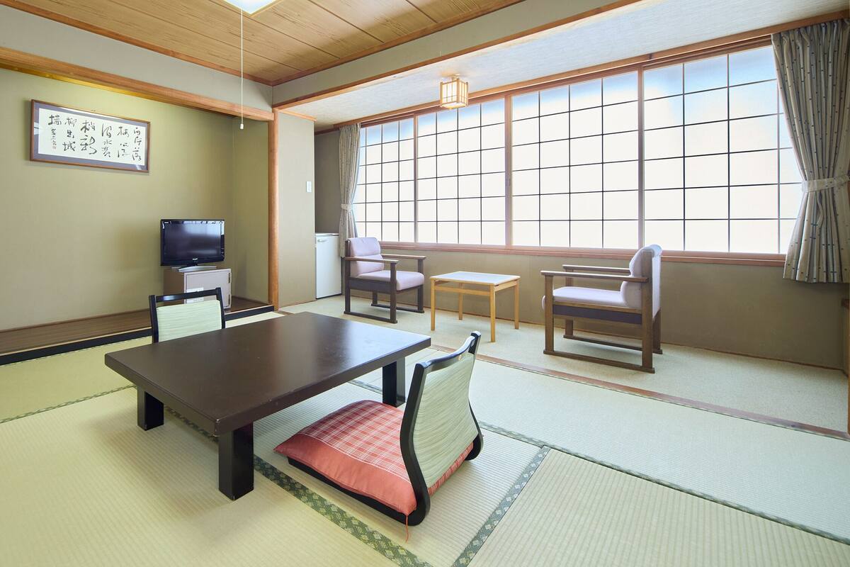 Ein traditioneller japanischer Raum mit Tatami-Böden, einem niedrigen Tisch, Stühlen, einem Fernseher und einem großen Fenster mit Vorhängen.