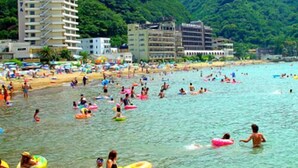 Ein Strand mit badenden Personen und Luftmatratzen, umgeben von Gebäuden und üppigen grünen Hügeln.