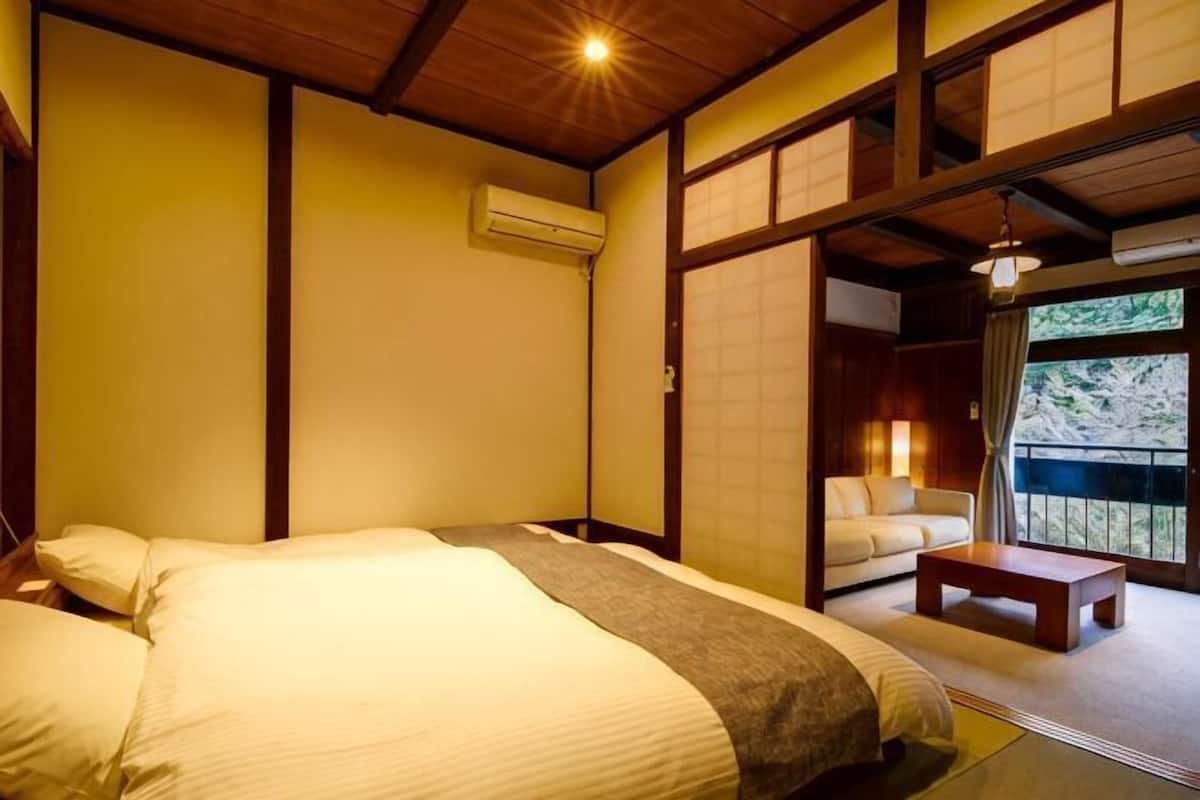 Ein traditioneller japanischer Raum mit Tatami-Boden, einem Bett mit weißer Bettwäsche, einer Holzbalkendecke und einer Schiebetür zu einem Außenbereich.