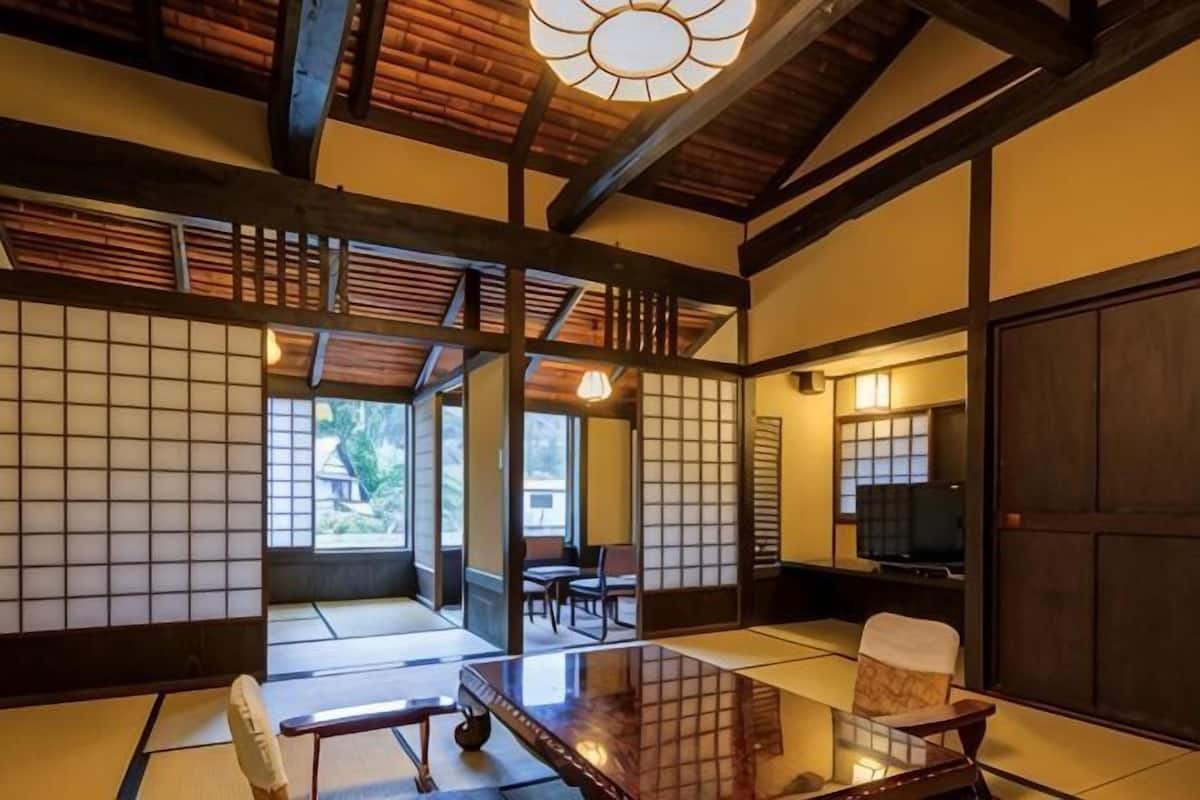 Ein traditioneller japanischer Raum mit Tatami-Bodenbelag, einem niedrigen Tisch mit Kissen, einem Fernseher und Schiebetüren.