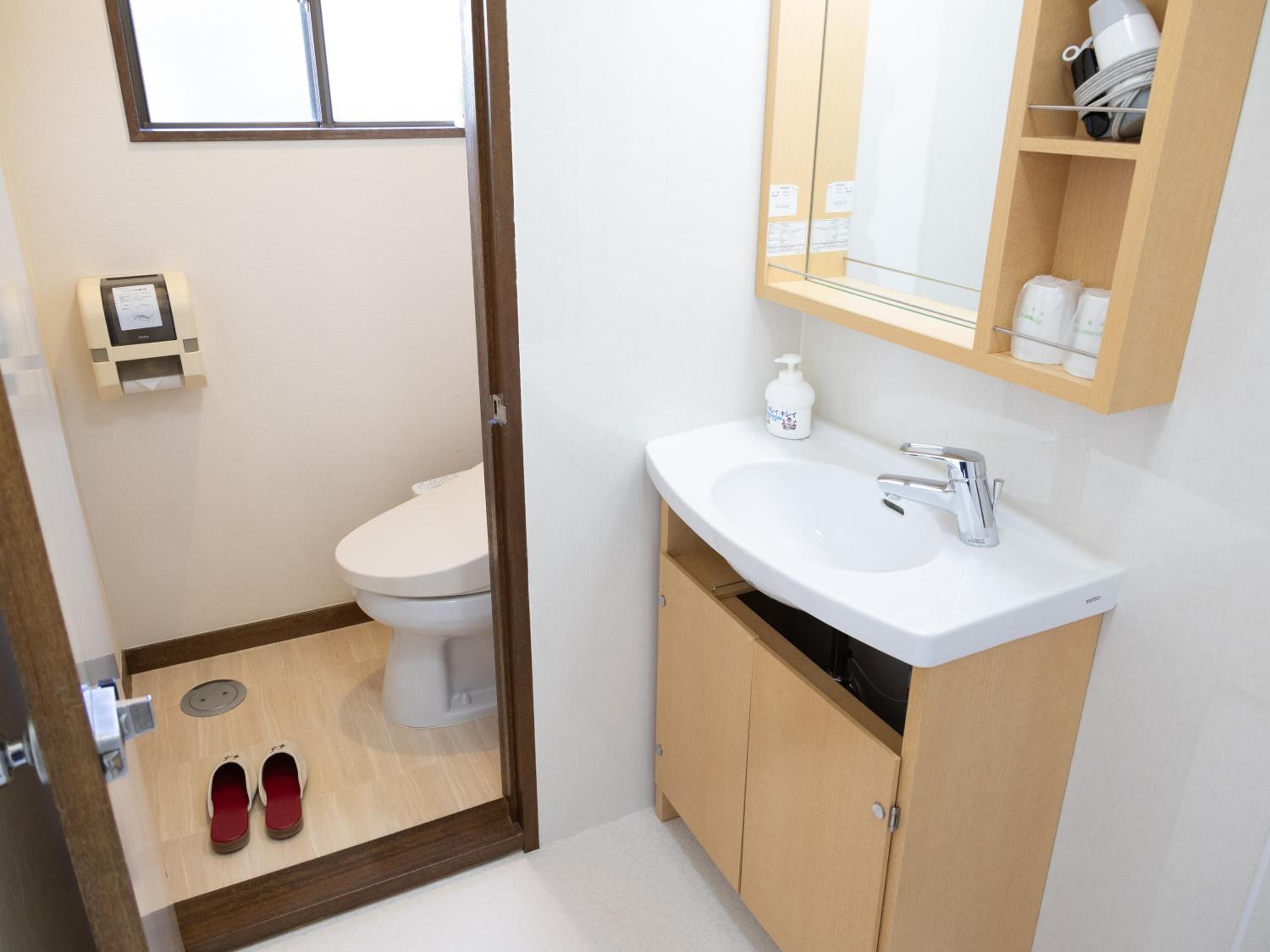 A bathroom with a toilet, a sink, and a mirror. There is a shelf with toiletries and a pair of shoes on the floor.