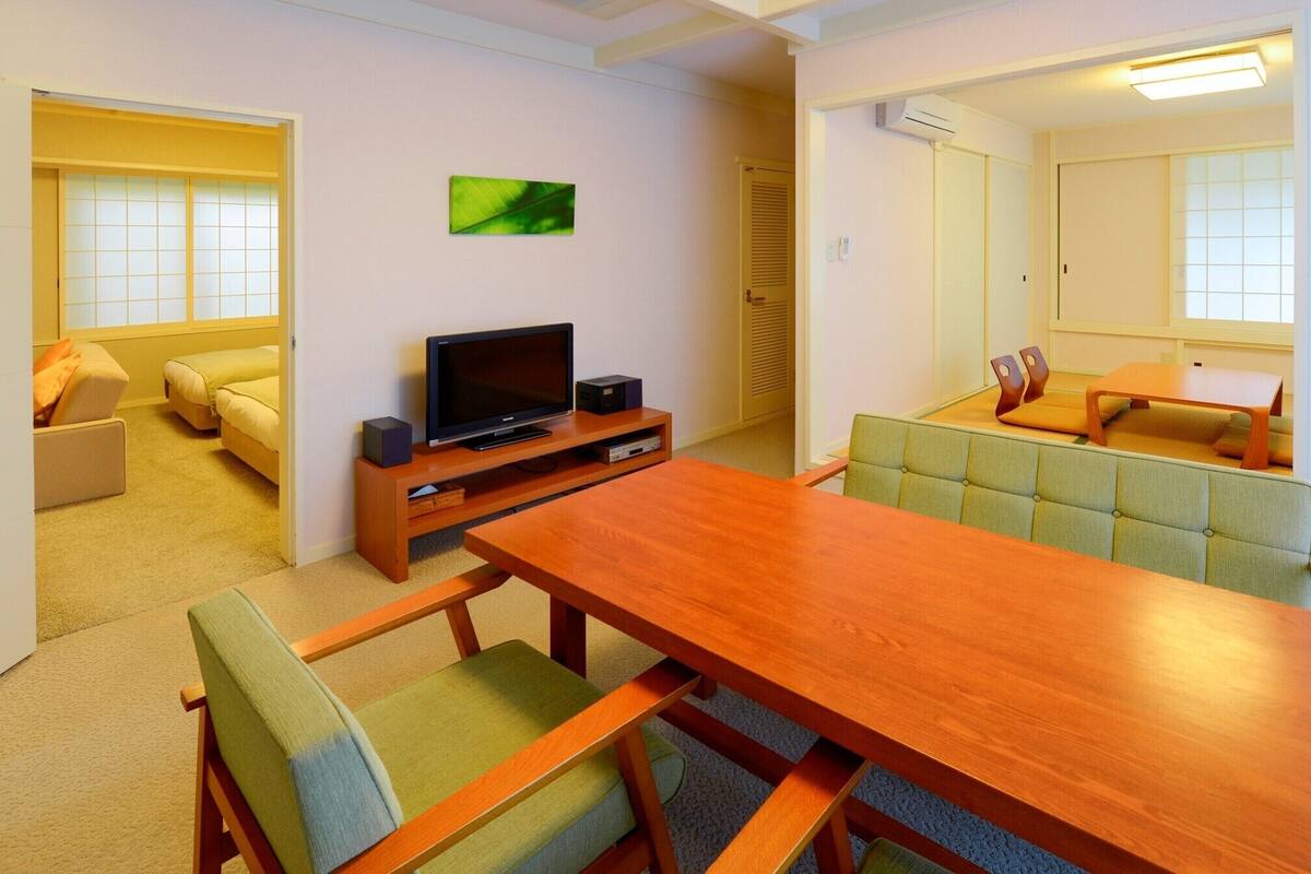 Ein modernes Wohnzimmer mit einem Holzessttisch, grünen Polsterstühlen, einem Flachbildfernseher auf einem Holzständer, einem Sofa und einem kleinen, grünen Gemälde an der Wand.