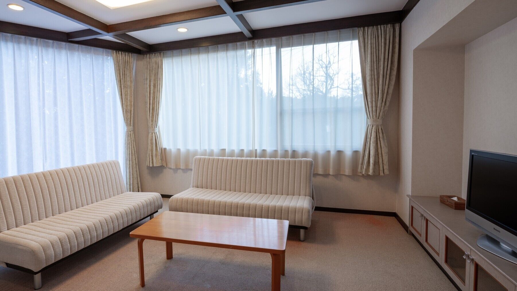 Ein Wohnzimmer mit einer Couch, einem zusätzlichen Sofa, einem Couchtisch, einem Fernseher und großen Fenstern mit Vorhängen.