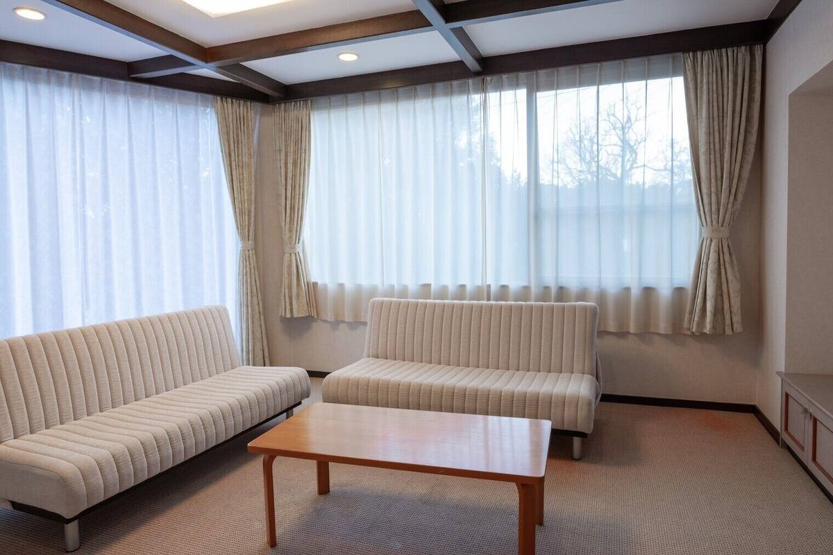 Ein Wohnzimmer mit einer Couch, einem zusätzlichen Sofa, einem Couchtisch, einem Fernseher und großen Fenstern mit Vorhängen.