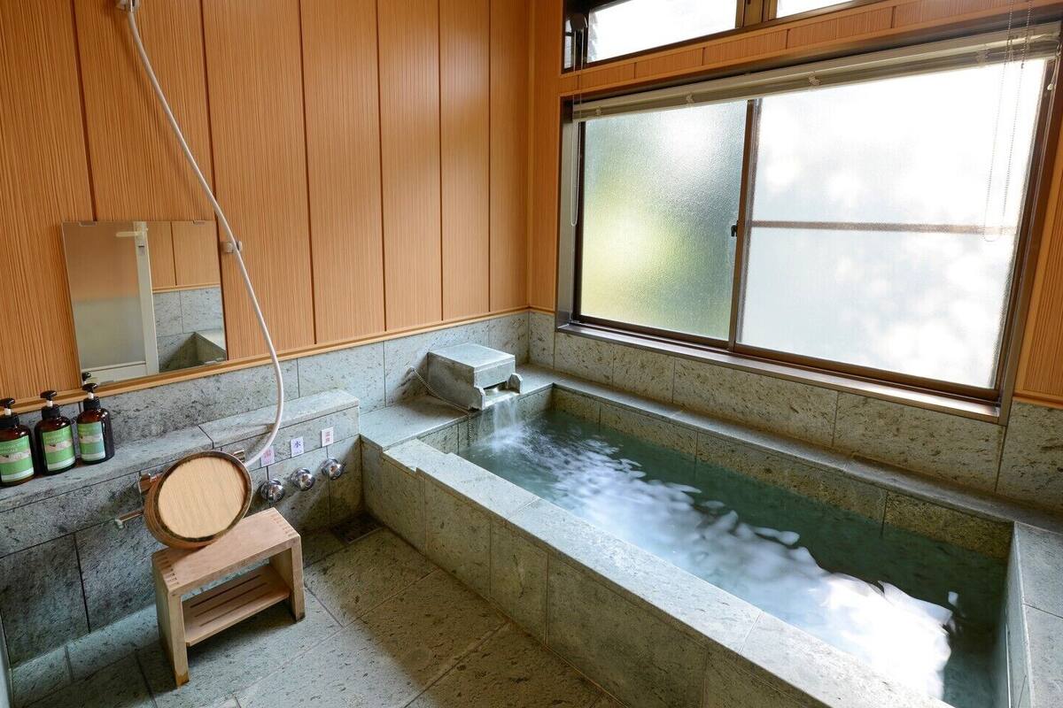 Ein traditionelles japanisches Onsen mit einer Holzbank, einer Steinsitzbadewanne und einem Fenster mit Blick auf den Himmel.