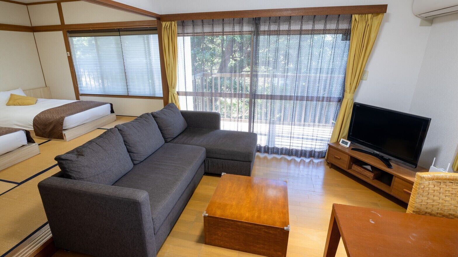 Uma sala de estar com sofá, televisão, mesa de centro de madeira e cama com travesseiro amarelo.