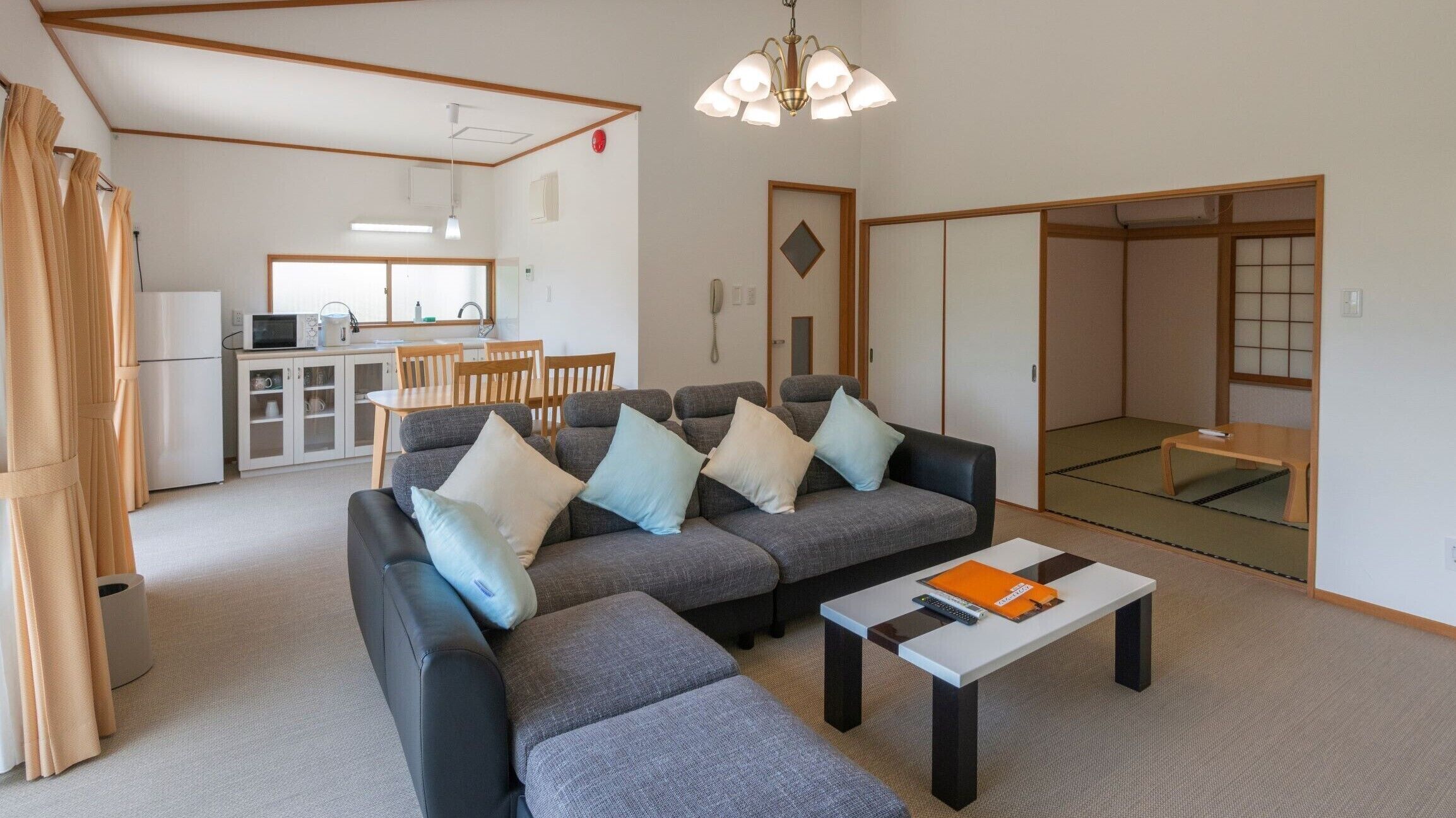 Uma sala de estar com sofá, apoio para os pés e mesa de centro, uma área de cozinha com geladeira, micro-ondas e pia, e uma porta japonesa tradicional deslizante.