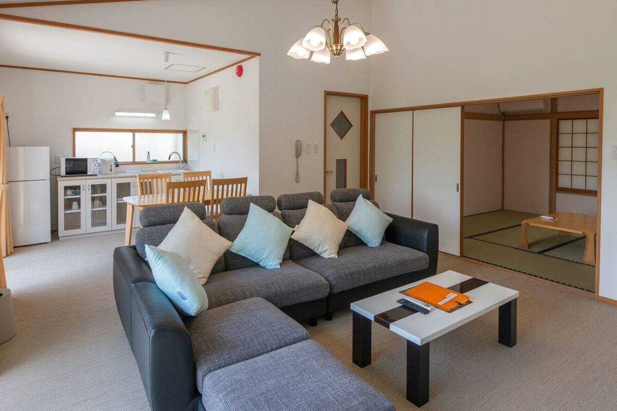Ein Wohnzimmer mit Sofa, Hocker und Couchtisch, ein Küchenbereich mit Kühlschrank, Mikrowelle und Spüle sowie eine traditionelle japanische Schiebetür.