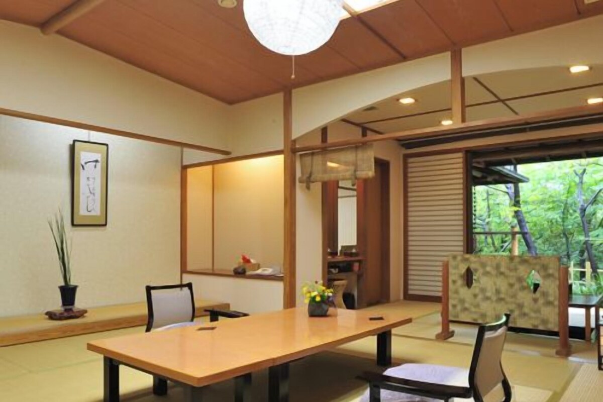 A traditional Japanese-style room with tatami flooring, a wooden table, chairs, a shoji screen, and a view of greenery outside.