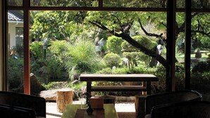 Vista a través de una ventana con un banco de madera y una mesa de piedra en primer plano, rodeado de abundante vegetación y árboles.