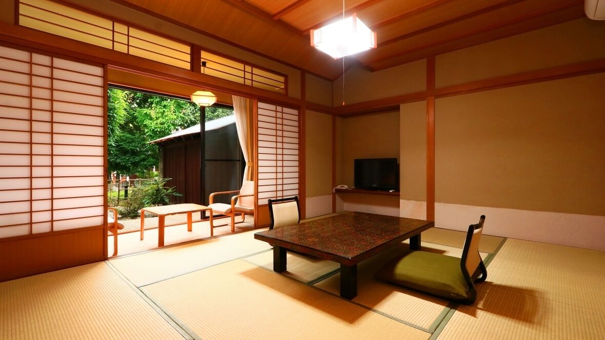 Una habitación japonesa tradicional con suelo de tatami, una mesa baja, sillas, un televisor y una puerta corredera que da a un espacio al aire libre.