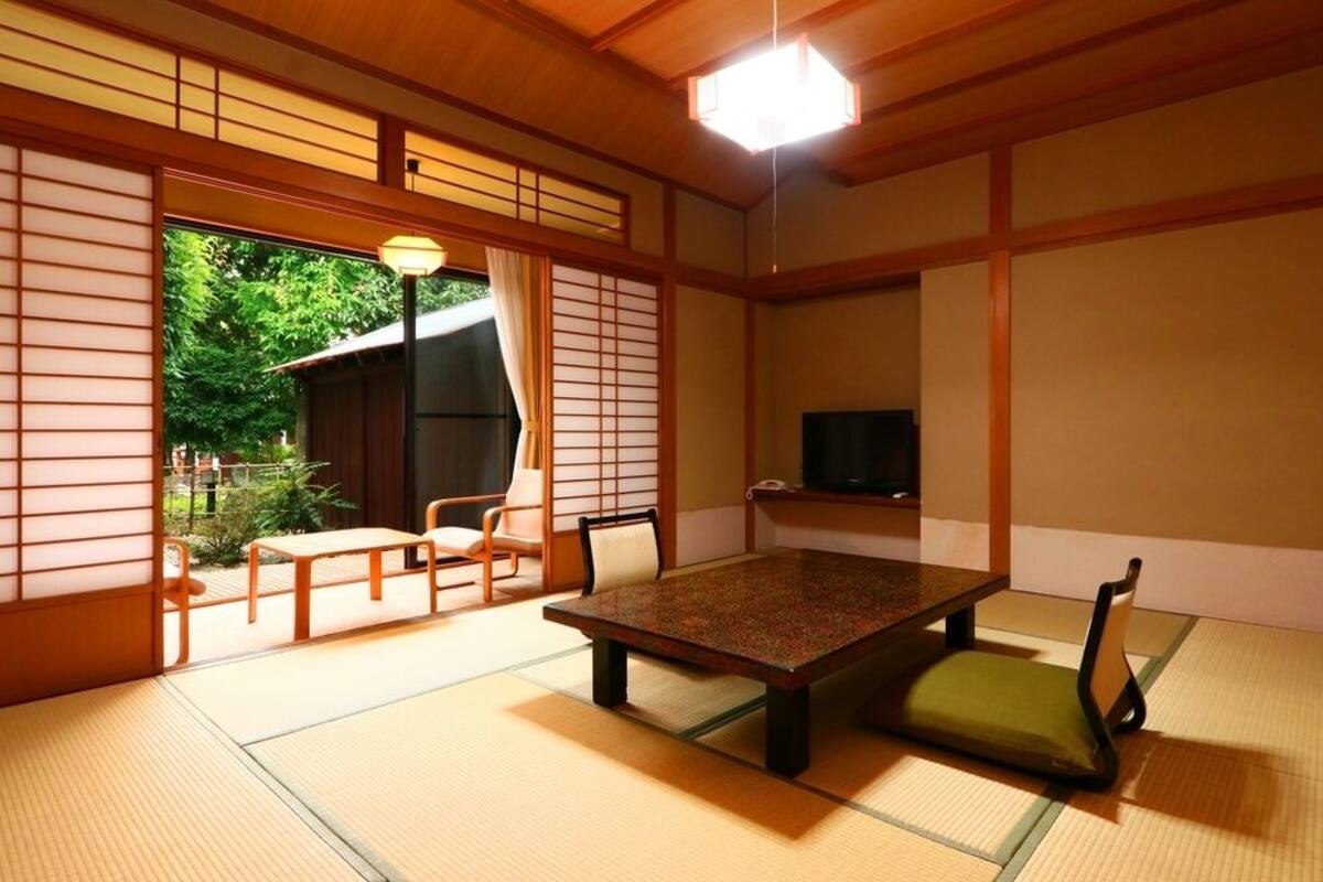 A traditional Japanese room with tatami flooring, a low table, chairs, a television, and a sliding door leading to an outdoor area.
