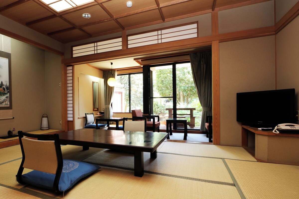 Una stanza tradizionale giapponese con pavimento in tatami, un tavolino, sedie, televisione e un'ampia finestra con vista sul verde.