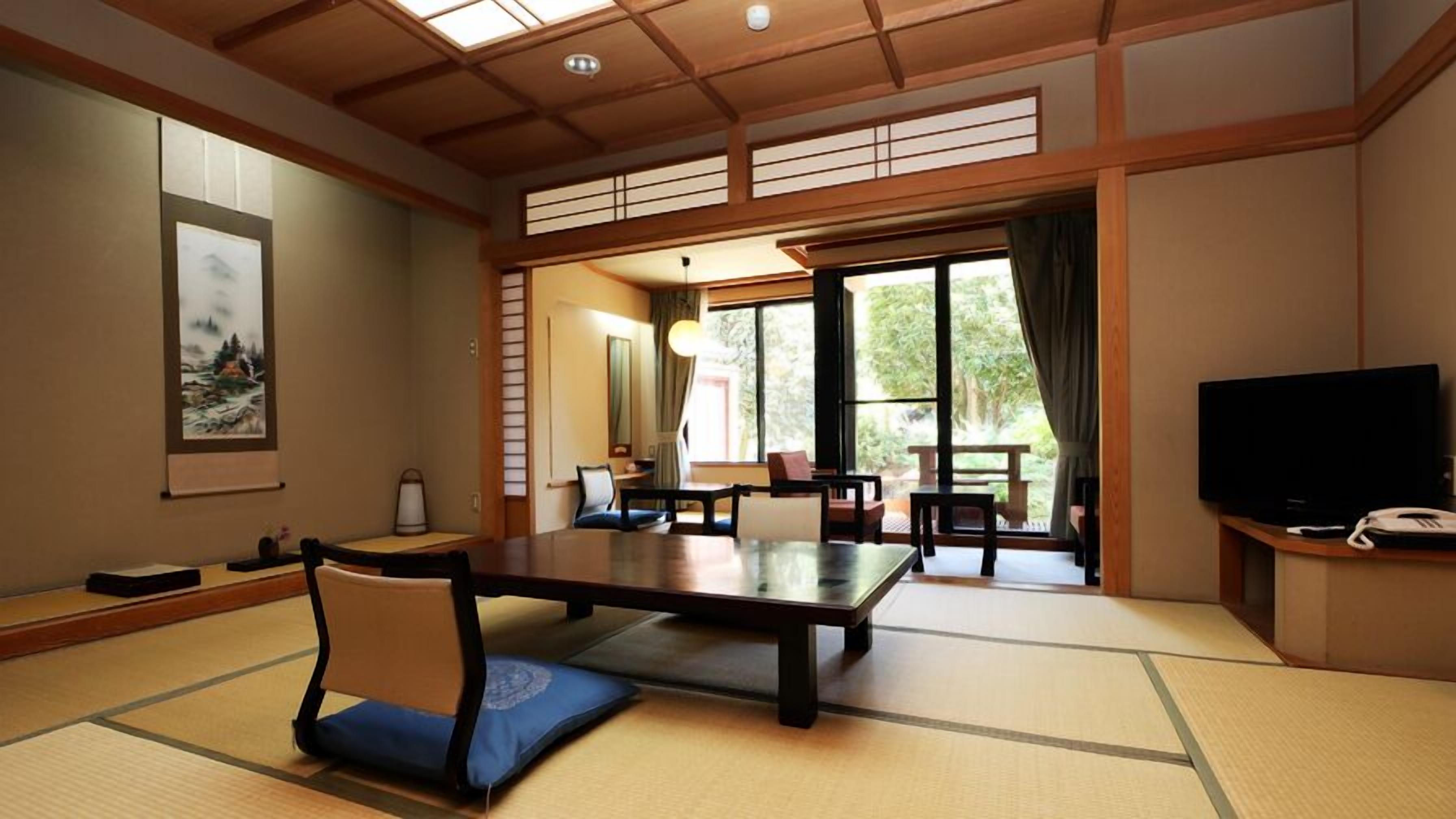 Una habitación tradicional japonesa con suelo de tatami, una mesa baja, sillas, un televisor y un ventanal con vistas al exterior.