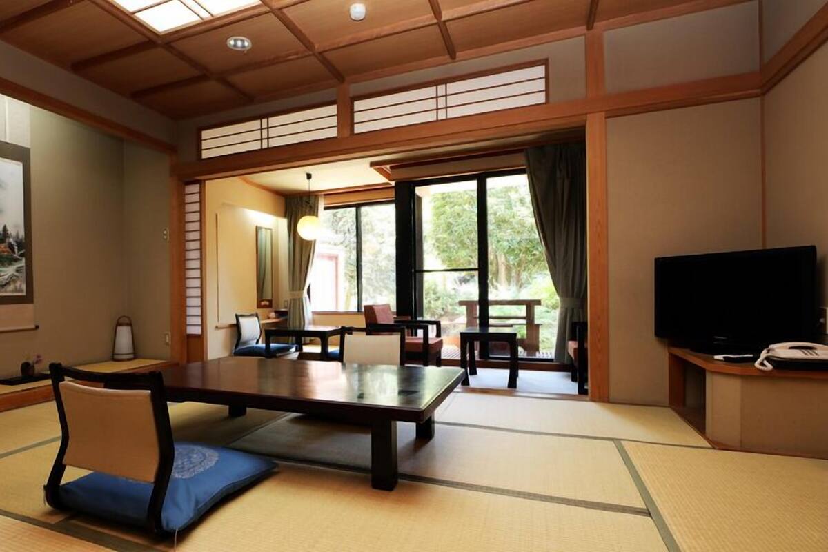 A traditional Japanese-style room with tatami flooring, a low table, chairs, a television, and a large window offering a view of the outdoors.