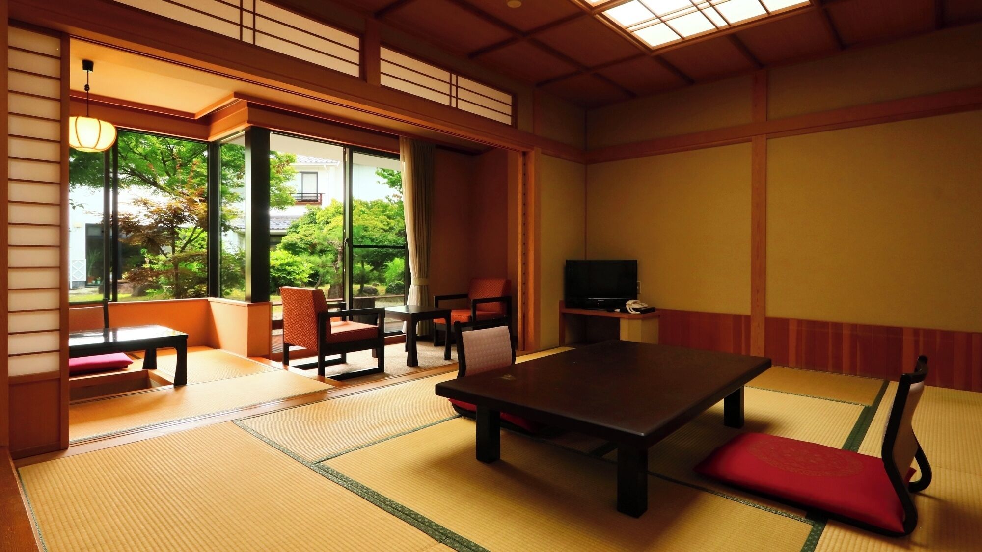 A traditional Japanese room with tatami flooring, a low table, chairs, a television, and large windows offering a view of greenery.