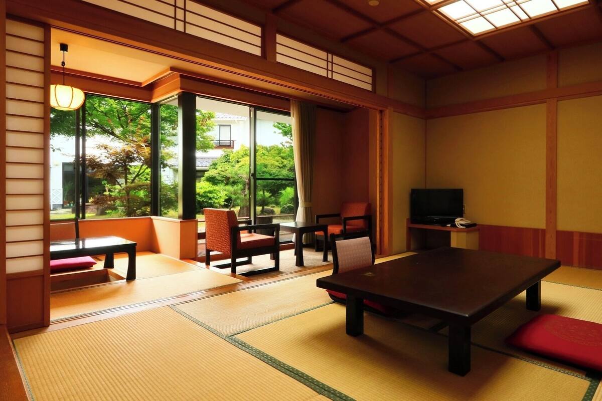 A traditional Japanese room with tatami flooring, a low table, chairs, a television, and large windows offering a view of greenery.