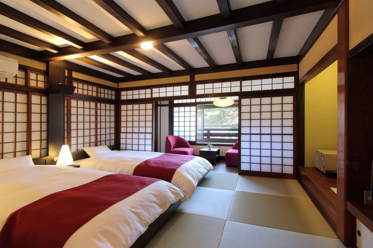 Une pièce de style japonais traditionnel, avec un sol recouvert de tatamis, deux lits avec une literie rouge et blanche, un bureau avec une lampe et une porte coulissante.
