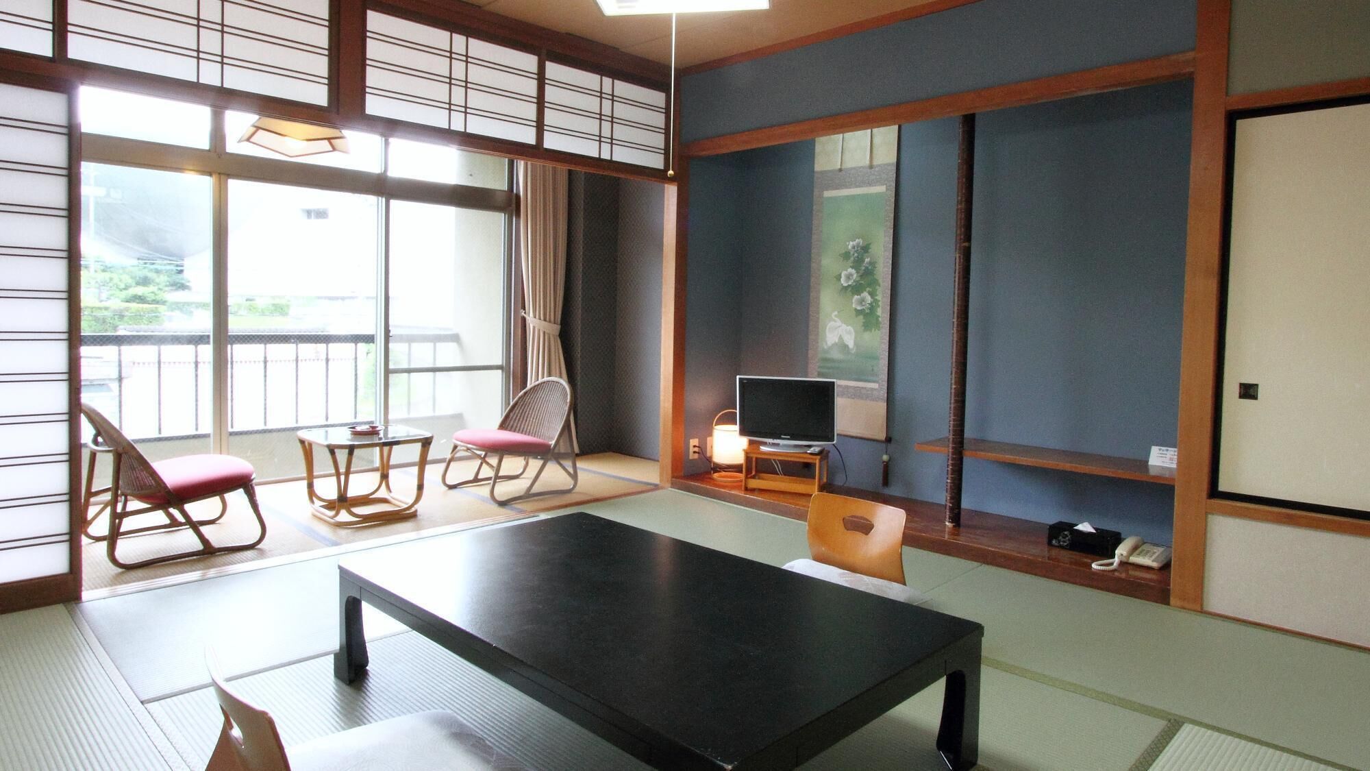 Una camera in stile tradizionale giapponese con pavimento in tatami, un tavolino, due sedie, una televisione e un'anta scorrevole.