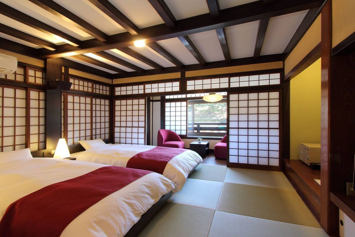 Une pièce de style japonais traditionnel, avec un sol recouvert de tatamis, deux lits avec une literie rouge et blanche, un bureau avec une lampe et une porte coulissante.