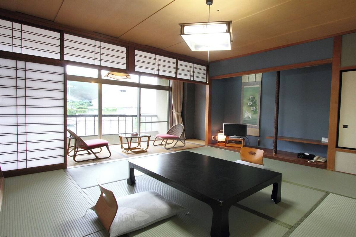 Une pièce de style japonais traditionnel, avec un sol recouvert de tatamis, une table basse, un téléviseur et une vue sur l’extérieur.