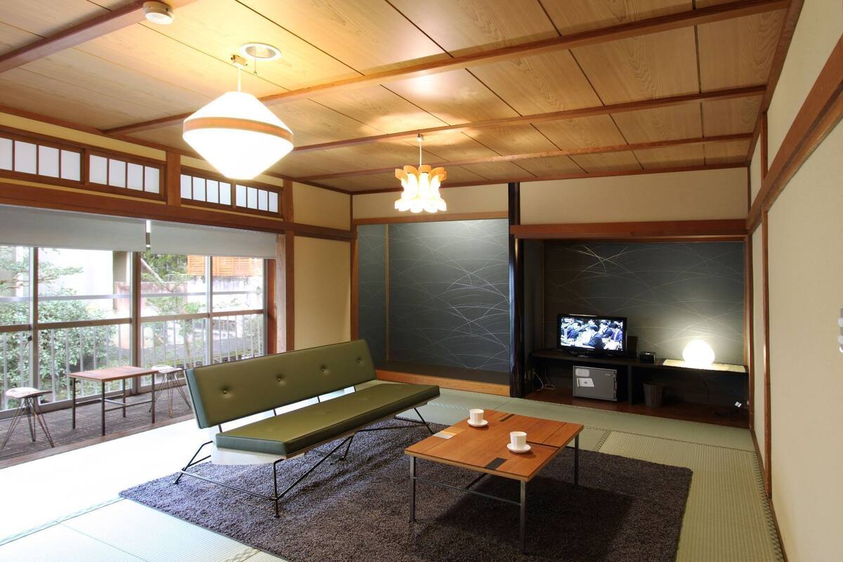 Une pièce de style japonais traditionnel, avec un sol recouvert de tatamis, un petit canapé vert, une table basse en bois, un téléviseur et une porte coulissante donnant sur un espace extérieur.
