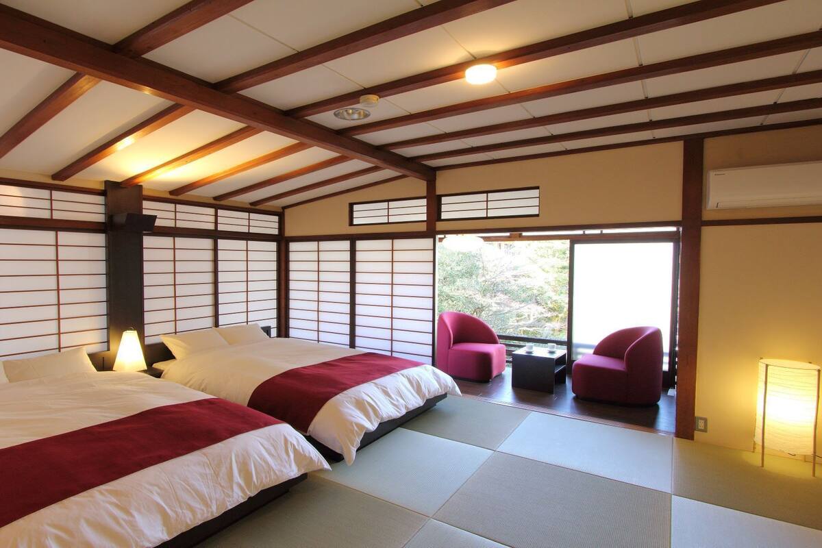 Une pièce de style japonais traditionnel, avec un sol recouvert de tatamis, deux lits avec une literie rouge, une porte coulissante et une vue sur la verdure à l’extérieur.